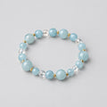Blue beaded bracelet on a white surface with green leaves and a candle in the background