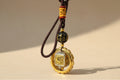 Decorative pendant with gold and black design on a brown cord against a gray background