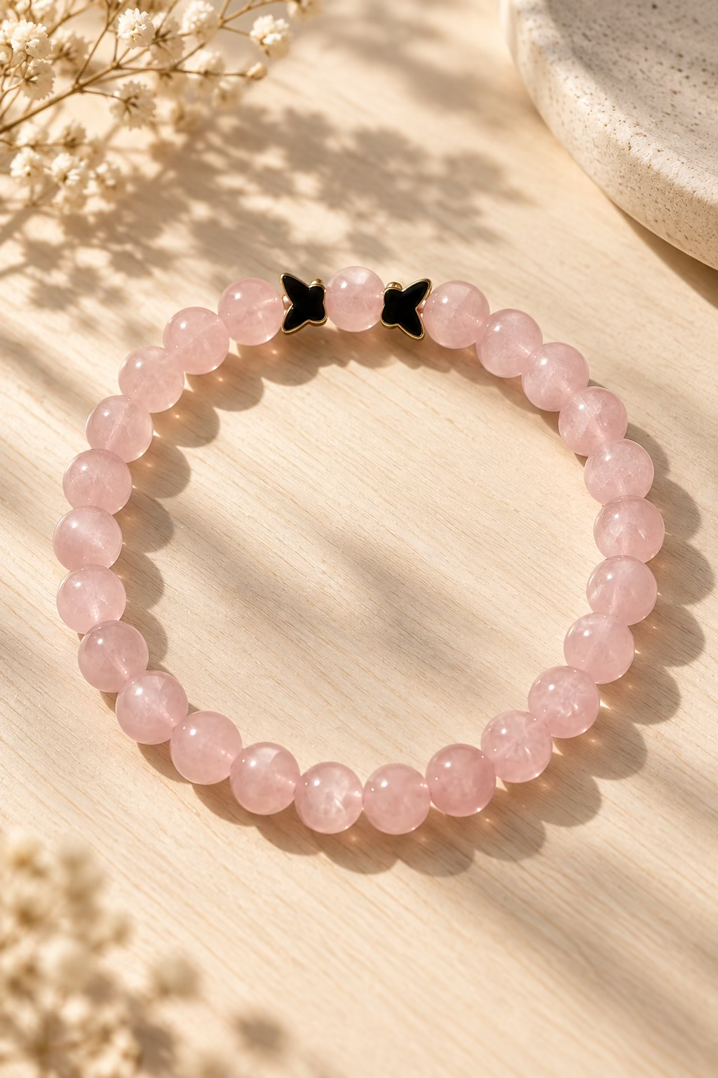 A pink stone beaded bracelet with a small black bow charm, displayed on a textured light gray surface.