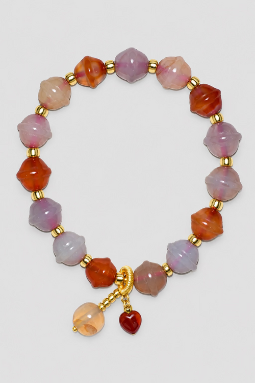 A beaded bracelet featuring faceted translucent beads in shades of pink, orange, and light purple, separated by small gold spacer beads, with a gold clasp and two dangling charms, one beige and one dark red, displayed on a textured light gray background.