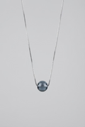 A minimalist necklace featuring a smooth, dark gray spherical bead pendant on a thin black chain, displayed against a textured gray background.