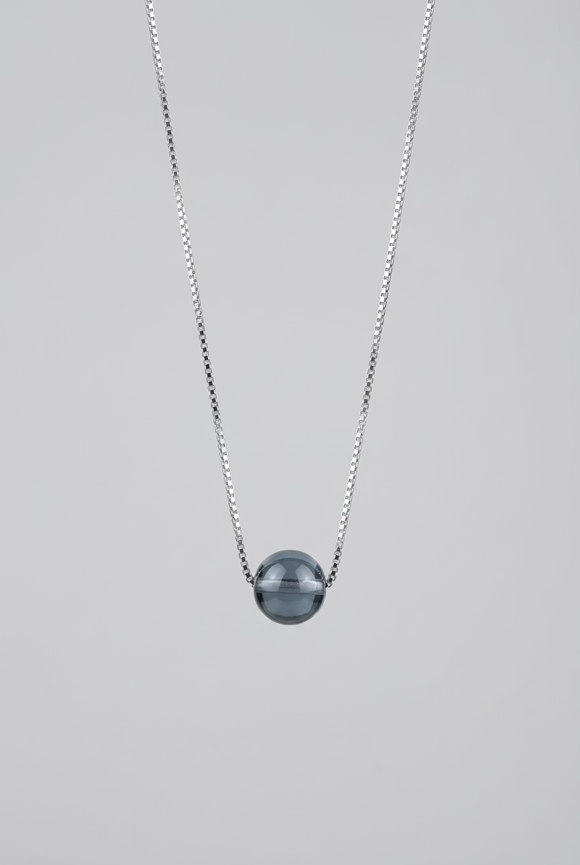 A minimalist necklace featuring a smooth, dark gray spherical bead pendant on a thin black chain, displayed against a textured gray background.