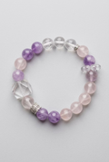 A beaded stretch bracelet featuring light purple amethyst beads, milky white quartz beads, a faceted white crystal, and a small silver-toned cross charm, displayed on a dark textured background.
