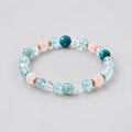 Multicolored beaded bracelet on a white surface with a small bowl and rolled fabric in the background.